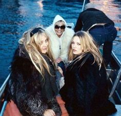 three women are sitting in a boat on the water, one is wearing a fur coat