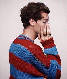 a young man covers his mouth with his hand while wearing a striped sweater and standing in front of a white wall