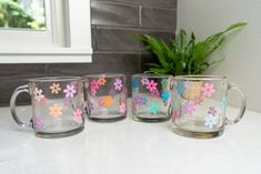 three glass mugs with flowers painted on them sitting next to a potted plant