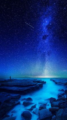 the night sky is filled with stars and clouds above rocks in the water, while a person stands at the edge of the ocean