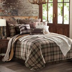 a bed with plaid comforter and pillows in front of a brick wall next to a window