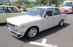 an old white car is parked in a parking lot with other cars and people standing around