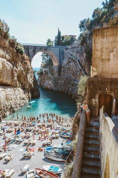 people are on the beach in front of an old stone bridge and some boats near by