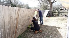 two men working on a wooden fence with one man reaching up to the fence and another person standing next to it