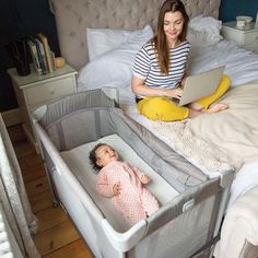 a woman sitting on a bed next to a baby in a crib with a laptop