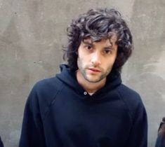 a man with curly hair wearing a black hoodie and looking at the camera while standing in front of a concrete wall