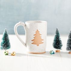 a coffee mug with christmas trees on it and other decorations around the cup, sitting on a white table