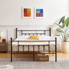 a bedroom with white walls and wood flooring has a black metal bed frame, two paintings on the wall