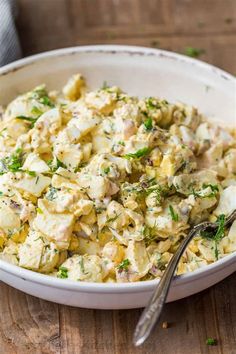 a white bowl filled with potato salad on top of a wooden table next to a fork