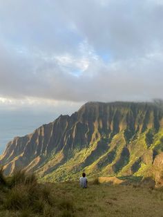 two people standing on top of a lush green hillside next to the ocean with mountains in the background