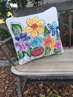 a painted pillow sitting on top of a wooden bench next to rocks and flowers in the background