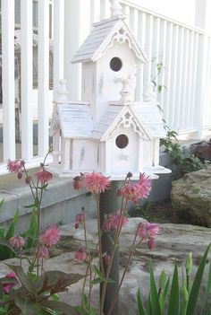 a white birdhouse sitting on top of a wooden pole next to flowers and rocks