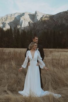 a bride and groom standing in tall grass with mountains in the background