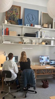 two people sitting at a desk in front of computer monitors and laptops, with bookshelves above them