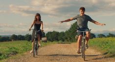 a man and woman riding bikes down a dirt road next to a lush green field