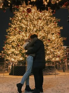 a man and woman embracing in front of a christmas tree at night with lights on it