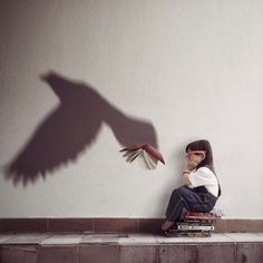 a woman sitting on top of a pile of books next to a shadow of a bird