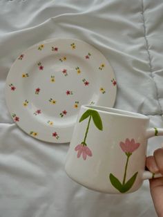 a hand holding a coffee cup next to a plate with flowers painted on the side