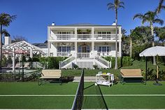 a tennis court with chairs and umbrellas in front of a large white house on the grass