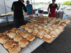 some people are standing by tables with food on it and there is a table full of baked goods