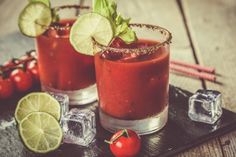 two glasses filled with red liquid and garnished with limes, tomatoes and ice cubes