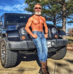 a shirtless man sitting on the hood of a black jeep in front of trees