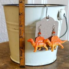 pair of dinosaur earrings with orange beads on hooks in front of bucket and measuring tape