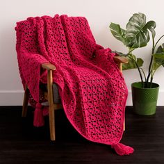 a pink crocheted blanket sitting on top of a wooden chair next to a potted plant