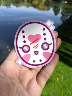 a hand holding a sticker with a cartoon character on it's face in front of a body of water