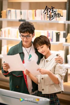 a man and woman standing next to each other in front of a book shelf holding papers