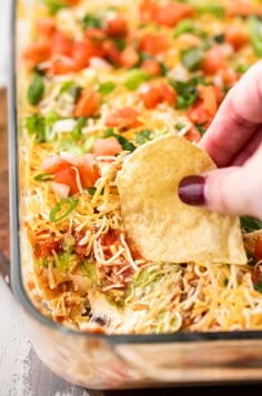 a hand dipping a tortilla chip into a casserole dish filled with shredded cheese and vegetables