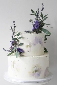 a three layer white cake with purple flowers and greenery on top, sitting on a plate