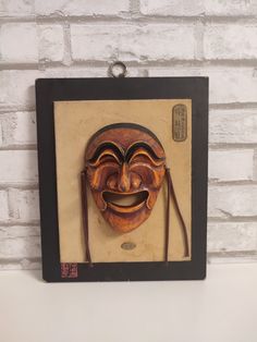 a wooden mask hanging on a brick wall with a black frame and brown leather strap