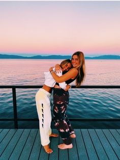 two women hugging each other while standing on a dock near the water at sunset or dawn