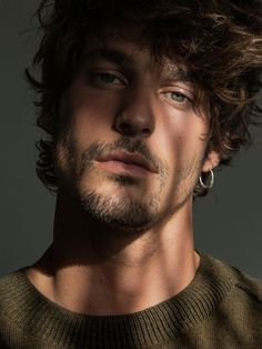 a man with curly hair and piercings looks at the camera while wearing a sweater