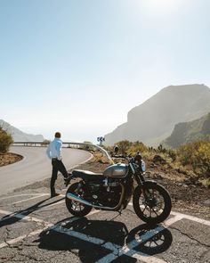 a man standing next to a motorcycle on the side of a road with mountains in the background