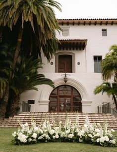 a large white building with palm trees and flowers in front