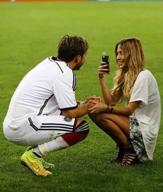 a man kneeling down next to a woman holding a cell phone on top of a field