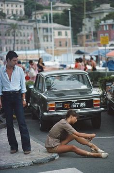 a man standing next to a woman sitting on the ground in front of a car