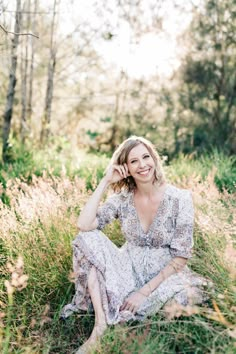 a woman is sitting in the grass with her hand on her head and smiling at the camera