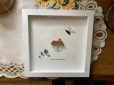 a white frame with a small house and butterflies in it on top of a table