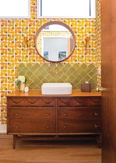 a bathroom with yellow and green wallpaper on the walls, a wooden dresser under a round mirror