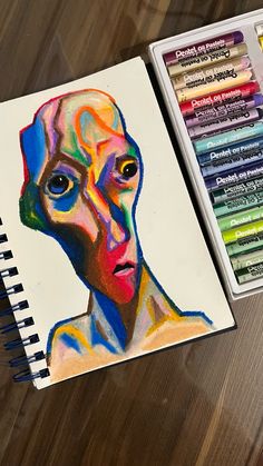 a drawing book with colored pencils next to it and a painting of a woman's face