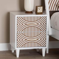 a nightstand with a clock on it next to a white bed and brown striped pillows