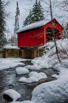 a red covered bridge over a small stream in the woods with snow on the ground