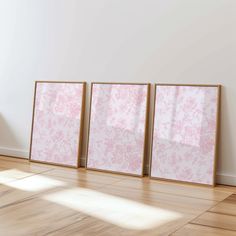 three frames on the floor in front of a white wall with pink floral designs and wooden floors