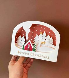 someone holding up a christmas card in front of a brown background with trees and houses