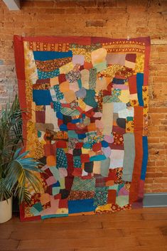 a large quilt hanging on the wall next to a potted plant and brick wall