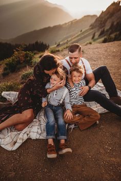 a family is sitting on a blanket in the mountains at sunset with their two children