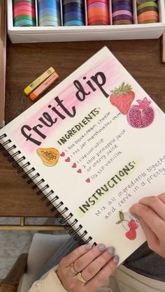 a woman is holding a notebook with fruit clip instructions on it and some crayons in the background